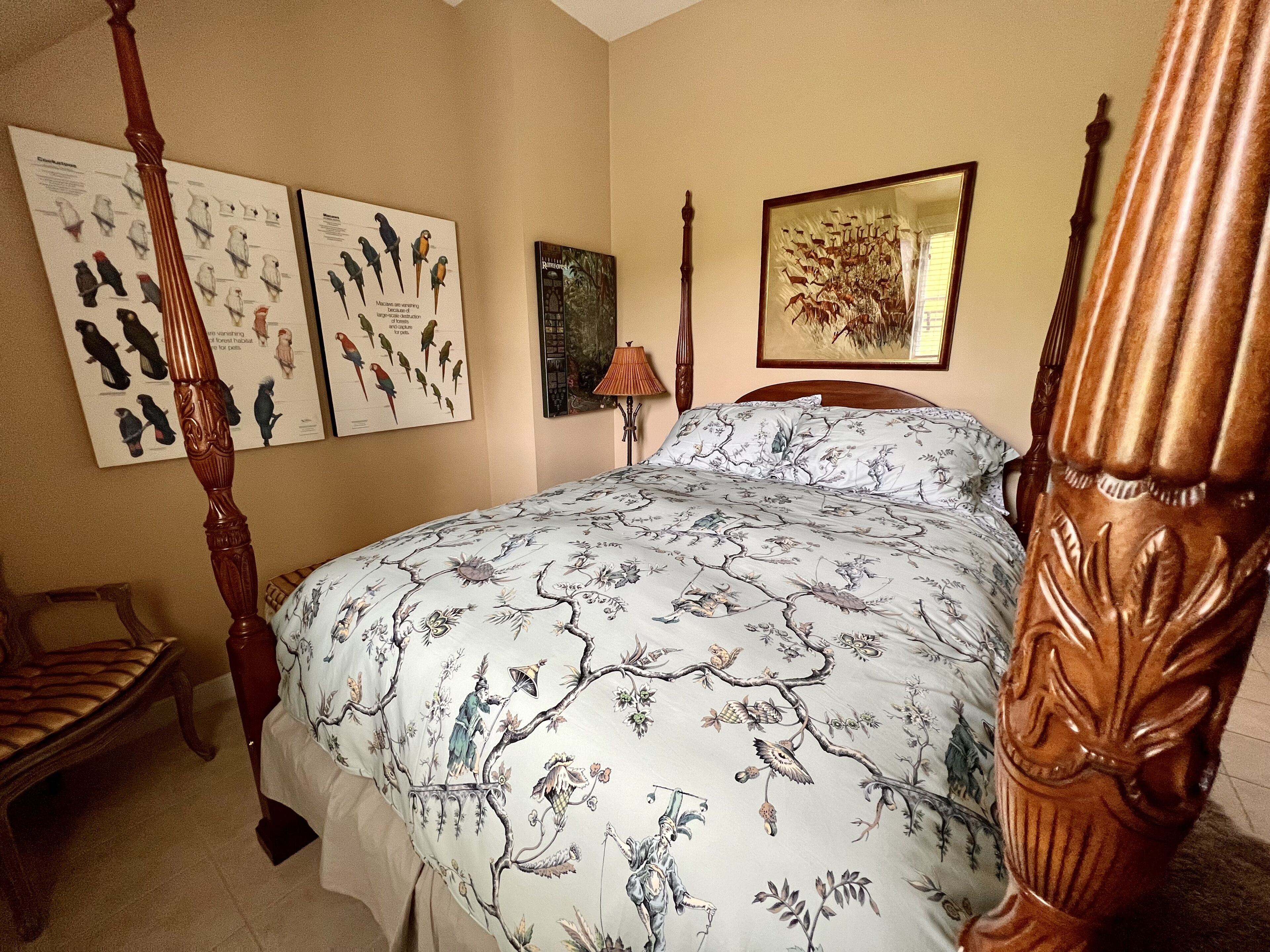 A four-poster bed with a floral bedspread, a wooden chair, and two wall-mounted bird illustrations.