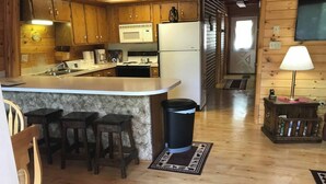 Una cocina con armarios de madera, microondas, refrigerador y fregadero. Hay taburetes de bar y un cubo de basura.