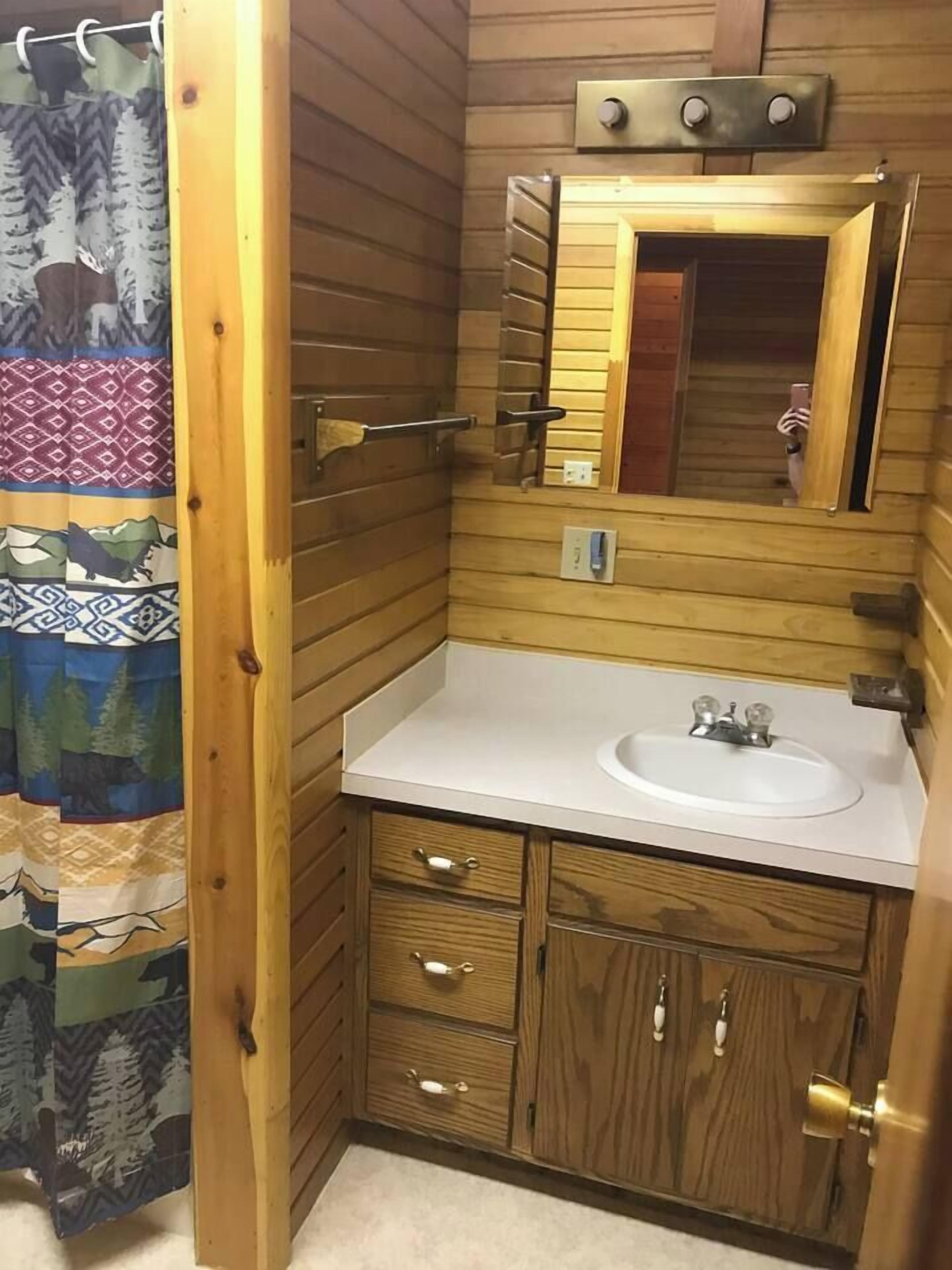 Ein Badezimmer mit Holzwandverkleidung, einem weißen Waschbecken, einem Spiegel und einem waschbärig gemusterten Duschvorhang.