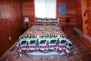 Ein Schlafzimmer in einer Blockhütte mit einem Bett, das ein Muster mit Bären und Elchen zeigt, einem Fenster mit Jalousien und einer wandmontierten Steckdose.
