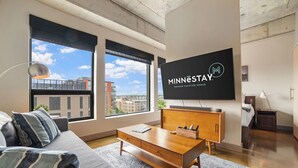 A modern living room with a large TV displaying 'Minnestay Premier Vacation Homes', a sofa, a coffee table, and large windows offering a city view. - MINNeSTAY* Sable 603 | North Loop | Target Center (Minneapolis)