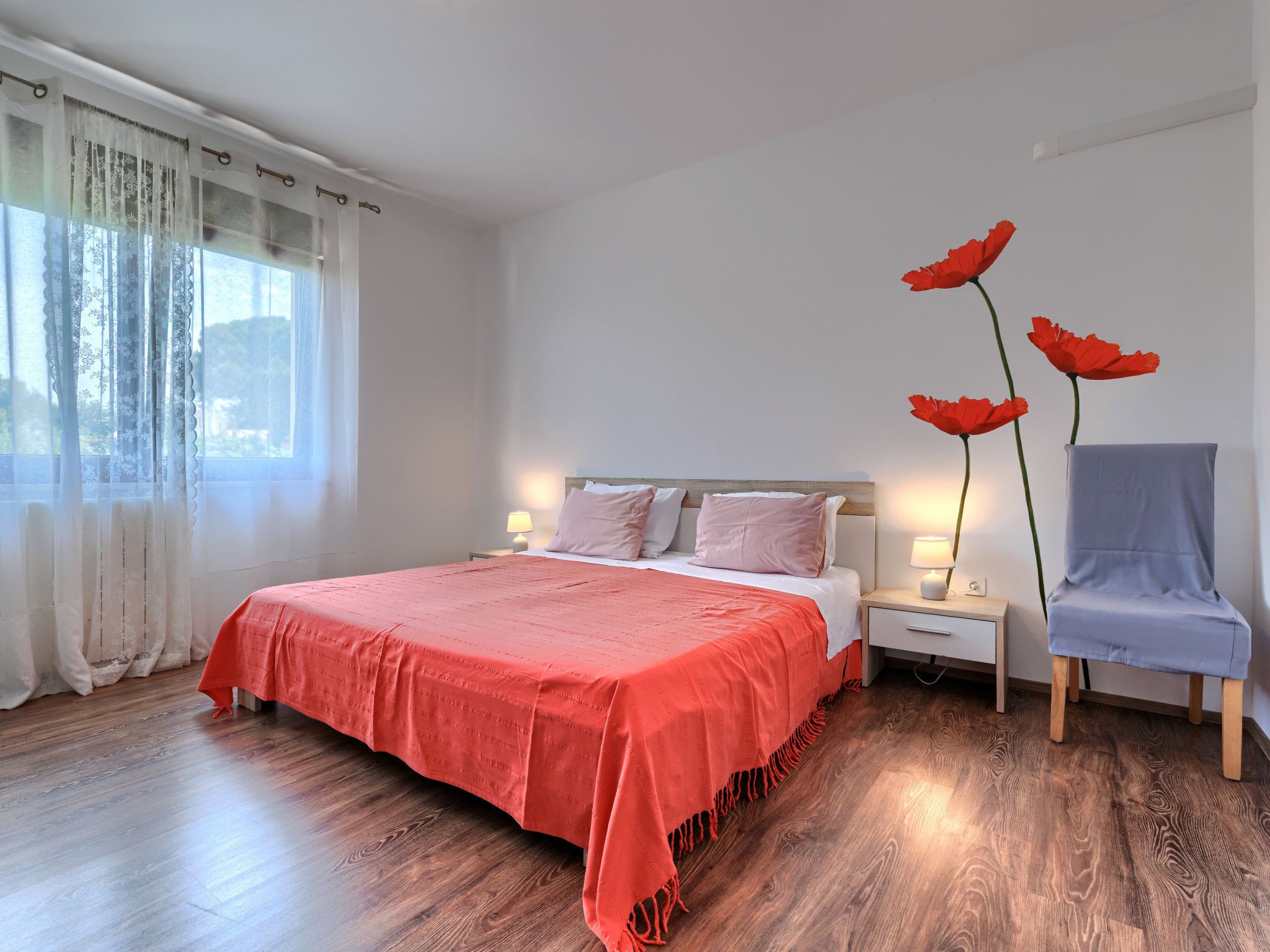 Une chambre à coucher avec un grand lit, des tables de chevet avec des lampes, un fauteuil et une fenêtre avec des rideaux légers.