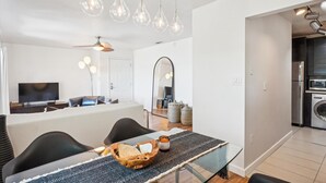 Una cocina moderna con electrodomésticos de acero inoxidable, un comedor con mesa y sillas de vidrio, y una sala de estar con televisor y asientos.