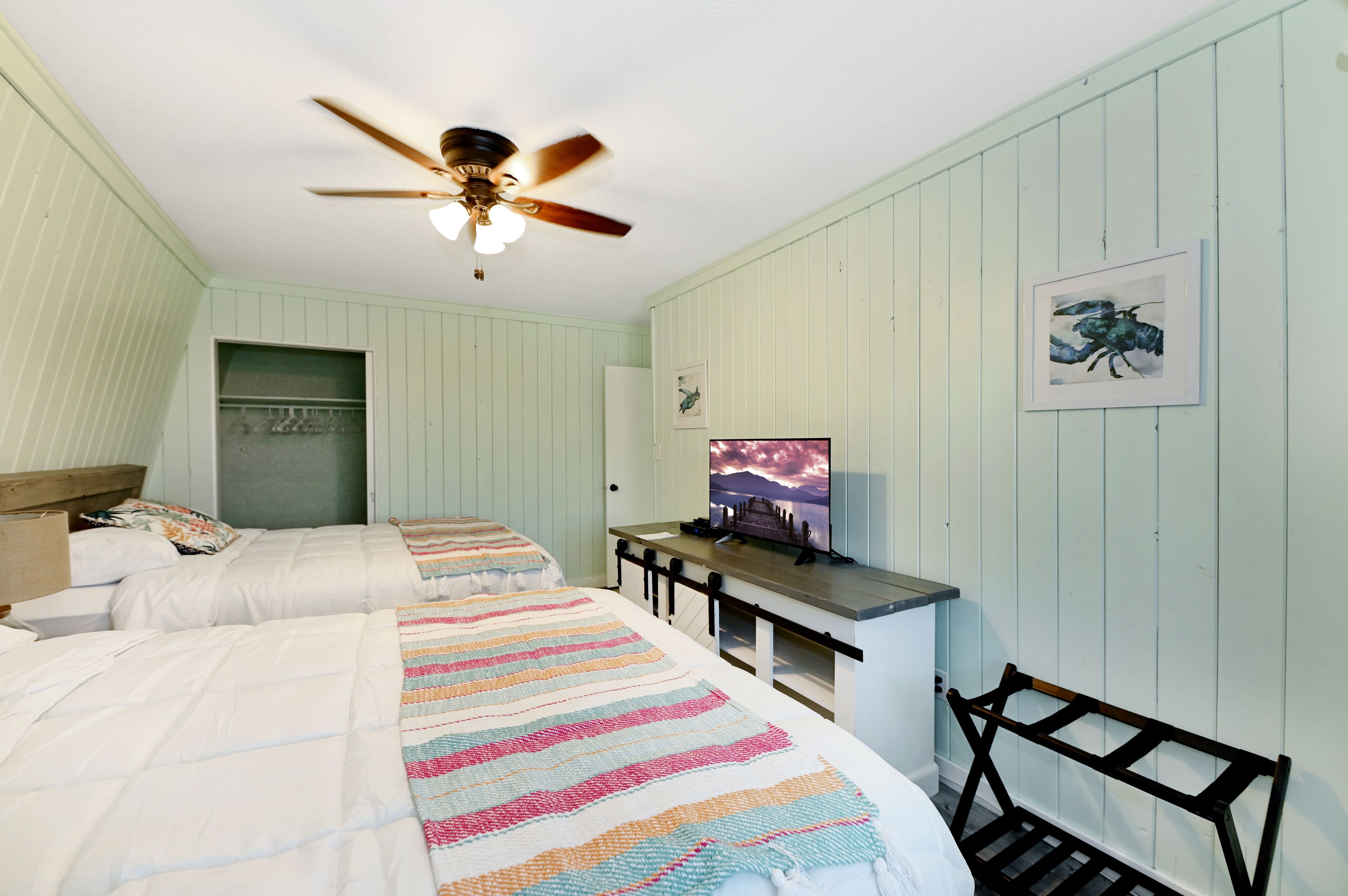 Une chambre avec deux lits, un ventilateur de plafond, une télévision et un tableau encadré au mur.