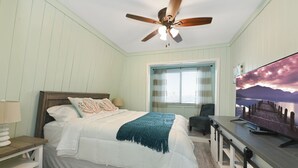 A bedroom with a bed, a desk, a chair, a ceiling fan, and a TV displaying a scenic image.