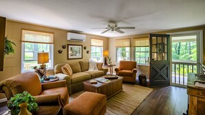 A cozy living room with a sofa, armchairs, a coffee table, and a ceiling fan. There are windows with blinds, a door leading outside, and a view of greenery.
