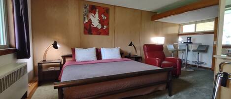Habitación de hotel con una cama grande, un sillón rojo, un escritorio con lámpara y un cuadro en la pared.
