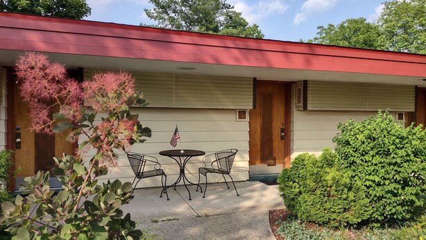 A motel with a red roof, a covered patio with two chairs and a small table, and a flagpole with an American flag. - King Bed - MCM Style- Backyard View (Spring Green)