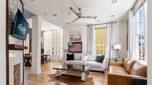 A spacious living room with a glass coffee table, leather sofas, a ceiling fan, and large windows with curtains. - Embrace Comfort and Convenience | Hibiscus House (Charleston)