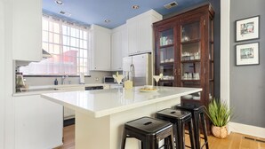 A modern kitchen with white cabinetry, a large island with bar stools, a wooden cabinet with glass doors, and a window with blinds. - Embrace Comfort and Convenience | Hibiscus House (Charleston)