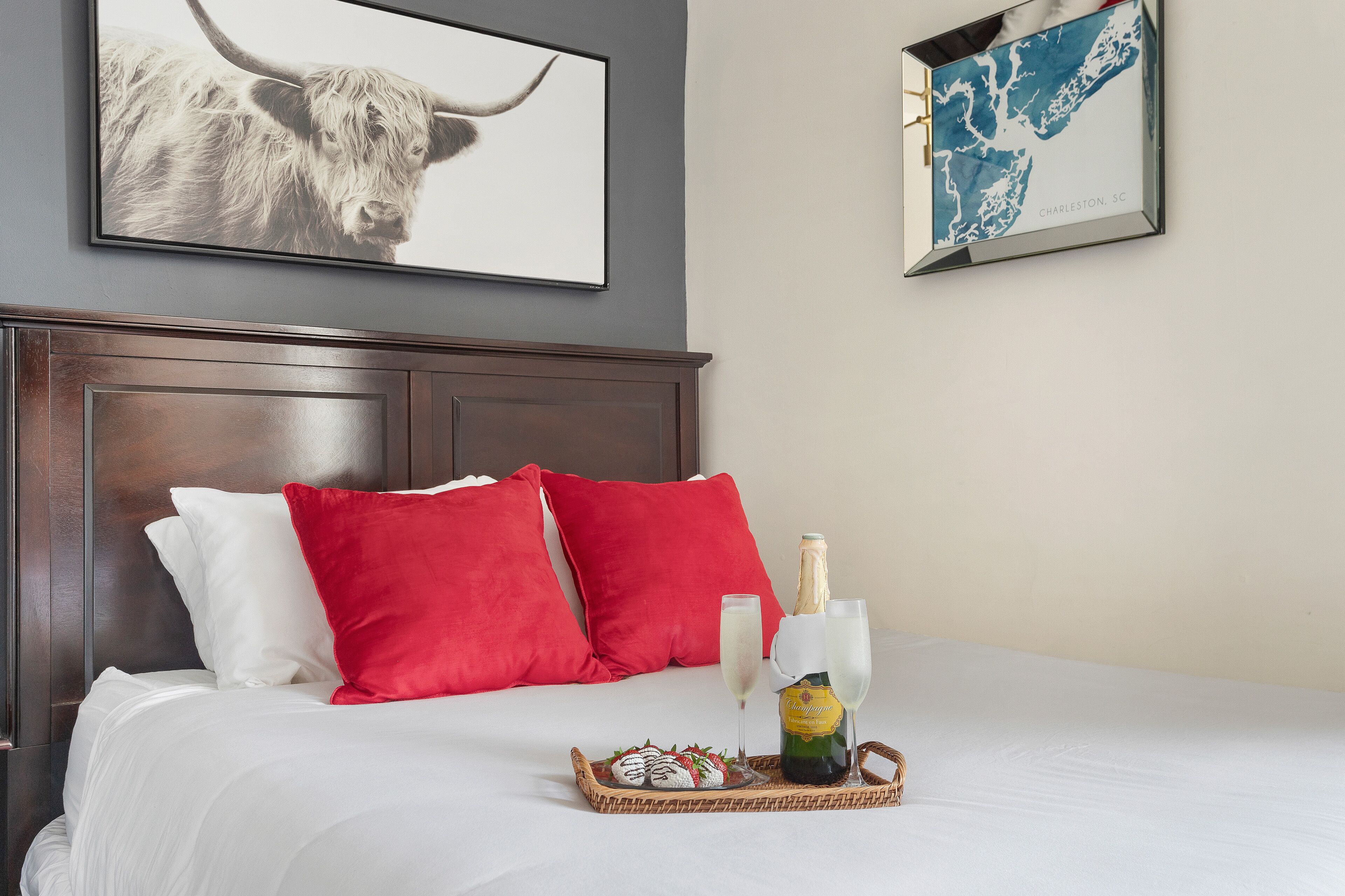 A neatly made bed with red and white pillows, a tray with a bottle and glasses, and two framed pictures on the wall.