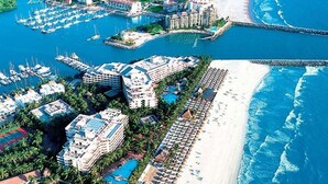 Vista aérea de un complejo turístico costero con varias edificaciones, un puerto deportivo y una playa.