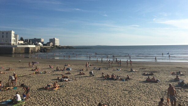 Een strand met mensen die zonnebaden en zwemmen, gebouwen op de achtergrond, en een heldere lucht.