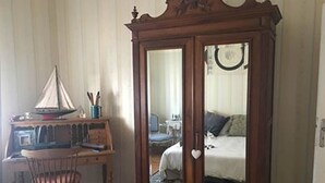 Een kamer met een houten garderobe, een klein bureau met een modelbootje, een stoel en een bed met kussens.