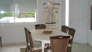 Een eetgedeelte met een ronde tafel, zes rieten stoelen, een kroonluchter en een raam met jaloezieën.