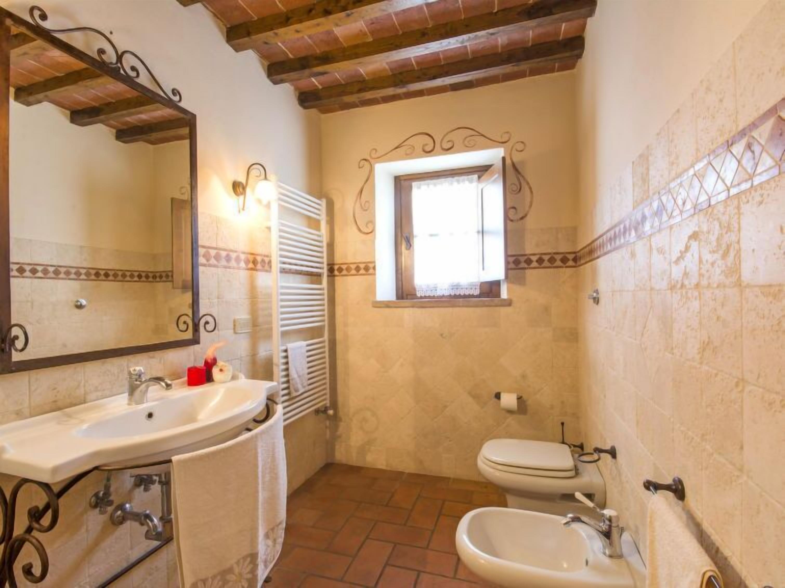 A bathroom with a sink, toilet, and bidet. It has a tiled floor and a window with a decorative frame.