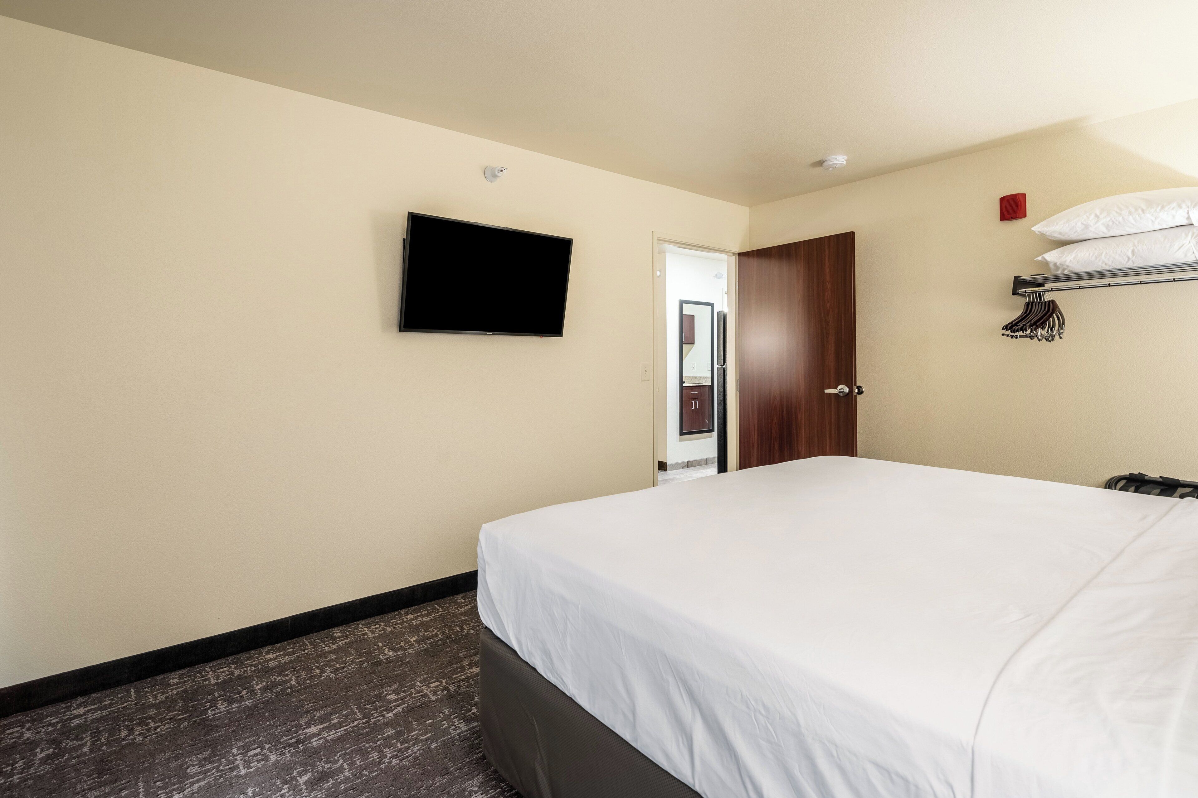 Ein Hotelzimmer mit einem Bett, einem an der Wand montierten Fernseher, einer Tür und einem Kleiderständer mit aufgehängten Kleidungsstücken.