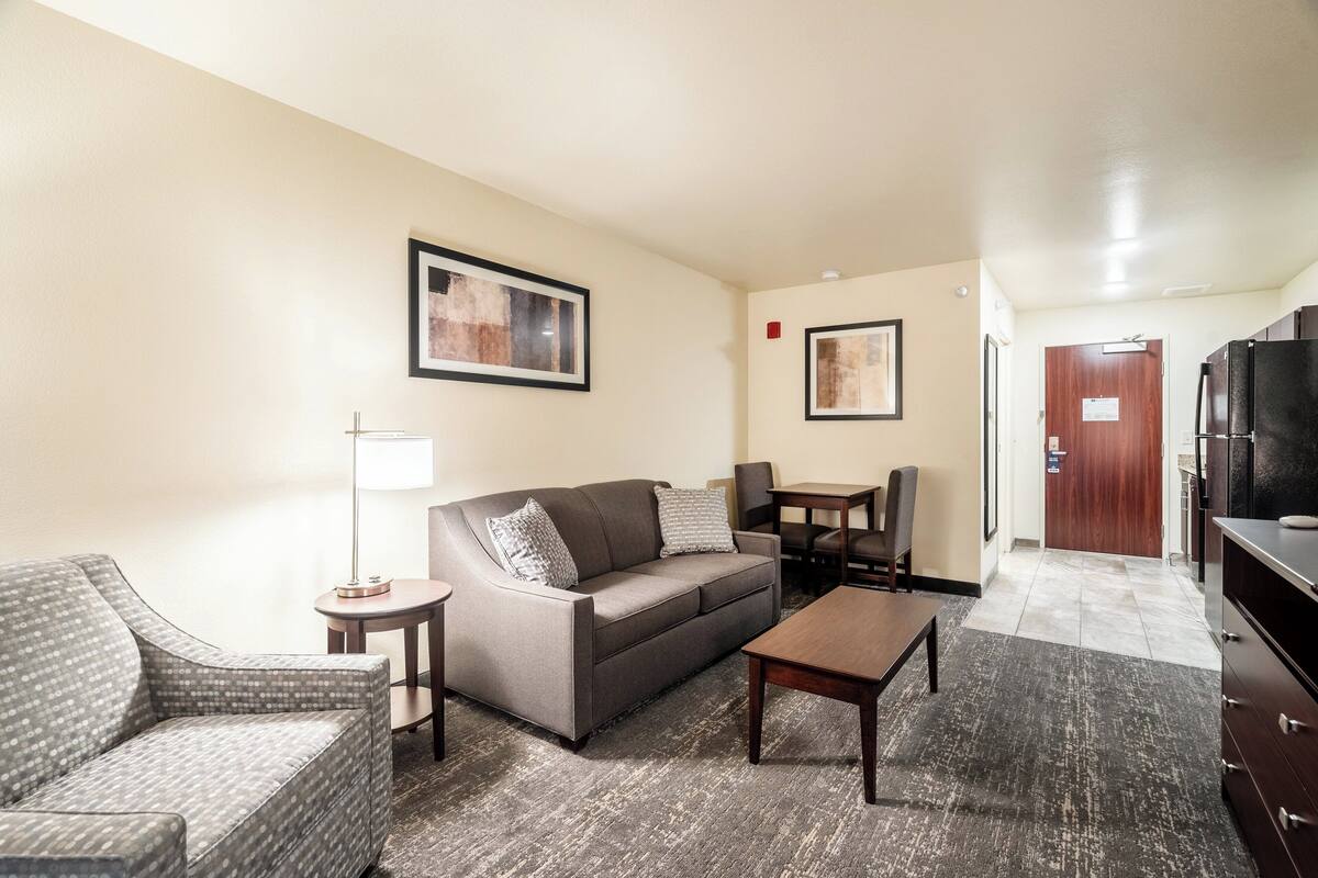 A hotel room with a sofa, armchair, coffee table, and a dining table with chairs. There are framed pictures on the walls and a door leading to the kitchen.