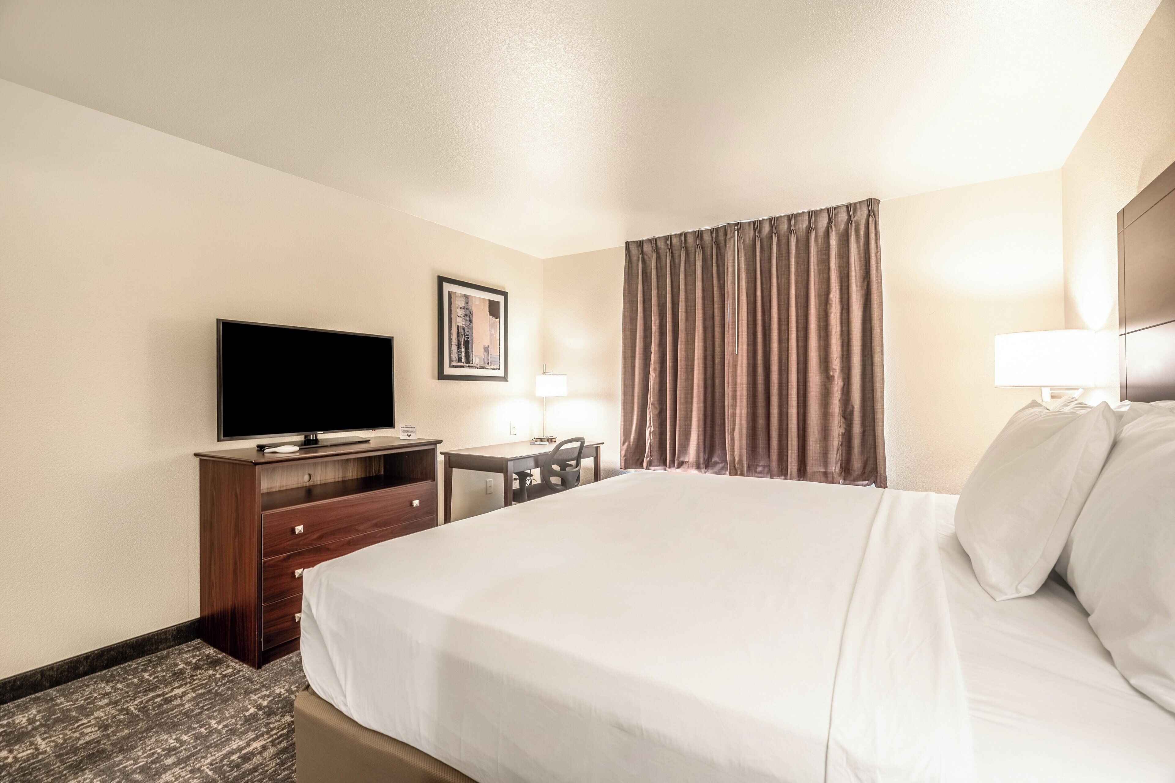 A hotel room with a large bed, a flat-screen TV, a desk with a chair, a lamp, and a window with curtains.