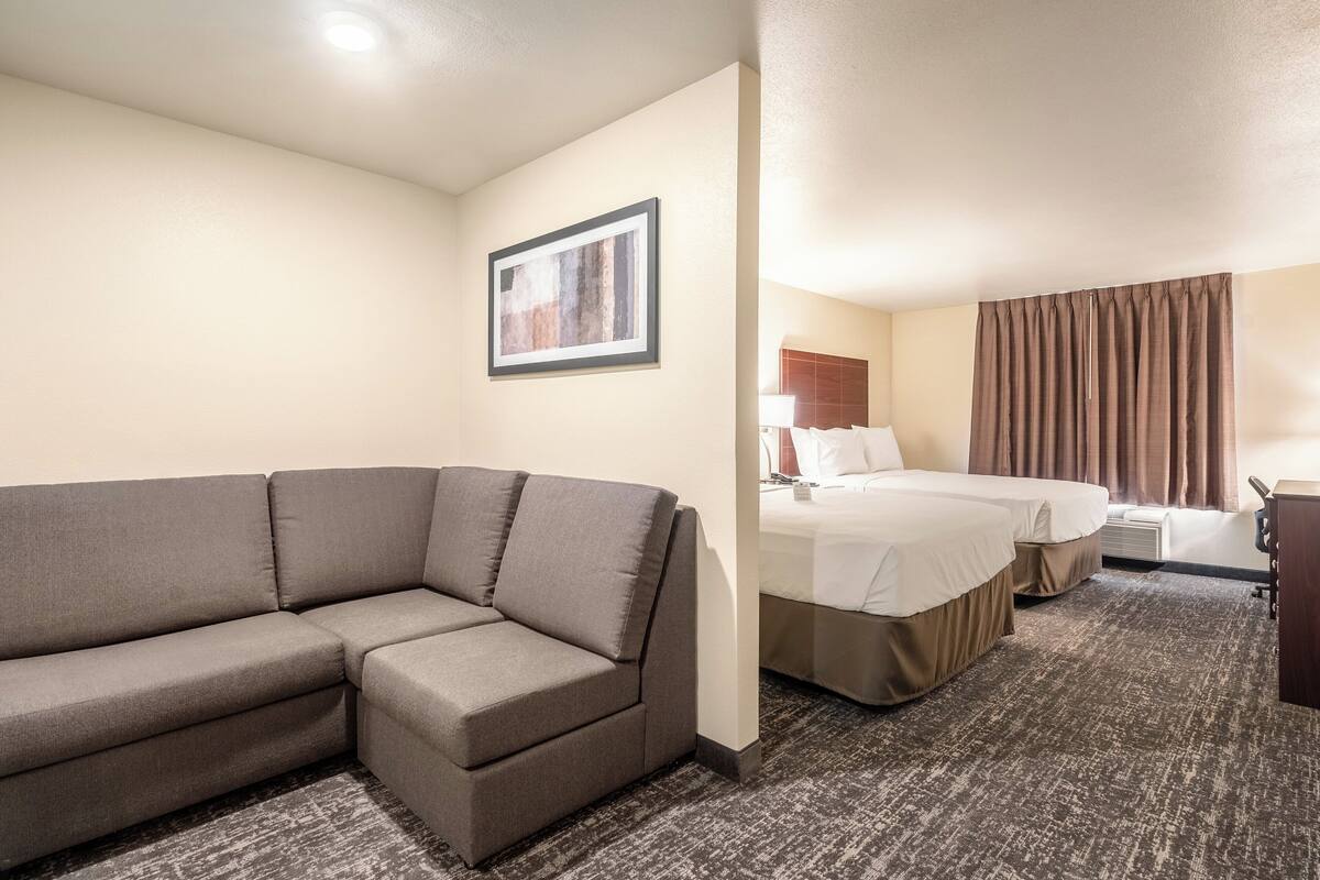 A hotel room with a gray sofa, two single beds, a desk, and a framed picture on the wall.