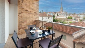 Un balcó amb taula i cadires, vista d'una població amb una torre d'església, i una paret de maó.