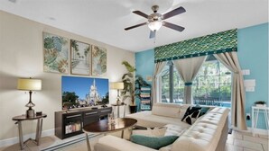 A living room with a sofa, coffee table, and a ceiling fan. There are framed pictures on the wall and a window with curtains. - Your Disney Family Vacation Resort Home With Private Pool/TikiBar (Davenport)