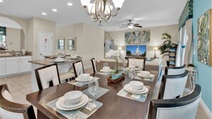A dining area with a wooden table set for four, surrounded by white chairs. A chandelier hangs from the ceiling, and there are framed pictures on the walls. - Your Disney Family Vacation Resort Home With Private Pool/TikiBar (Davenport)