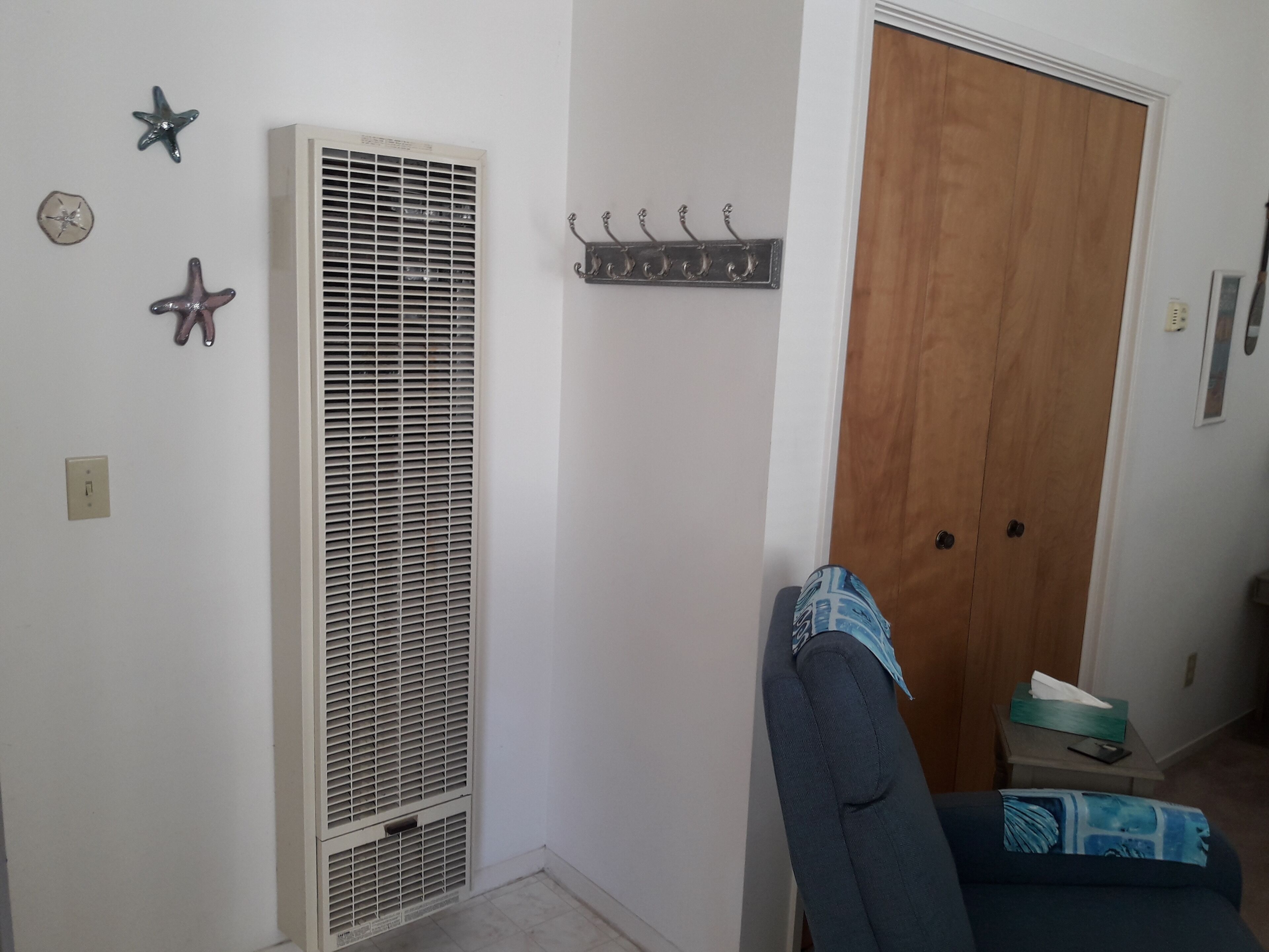 A room with a white wall, a vent, a wooden door, a chair, and a small table with a tissue box.