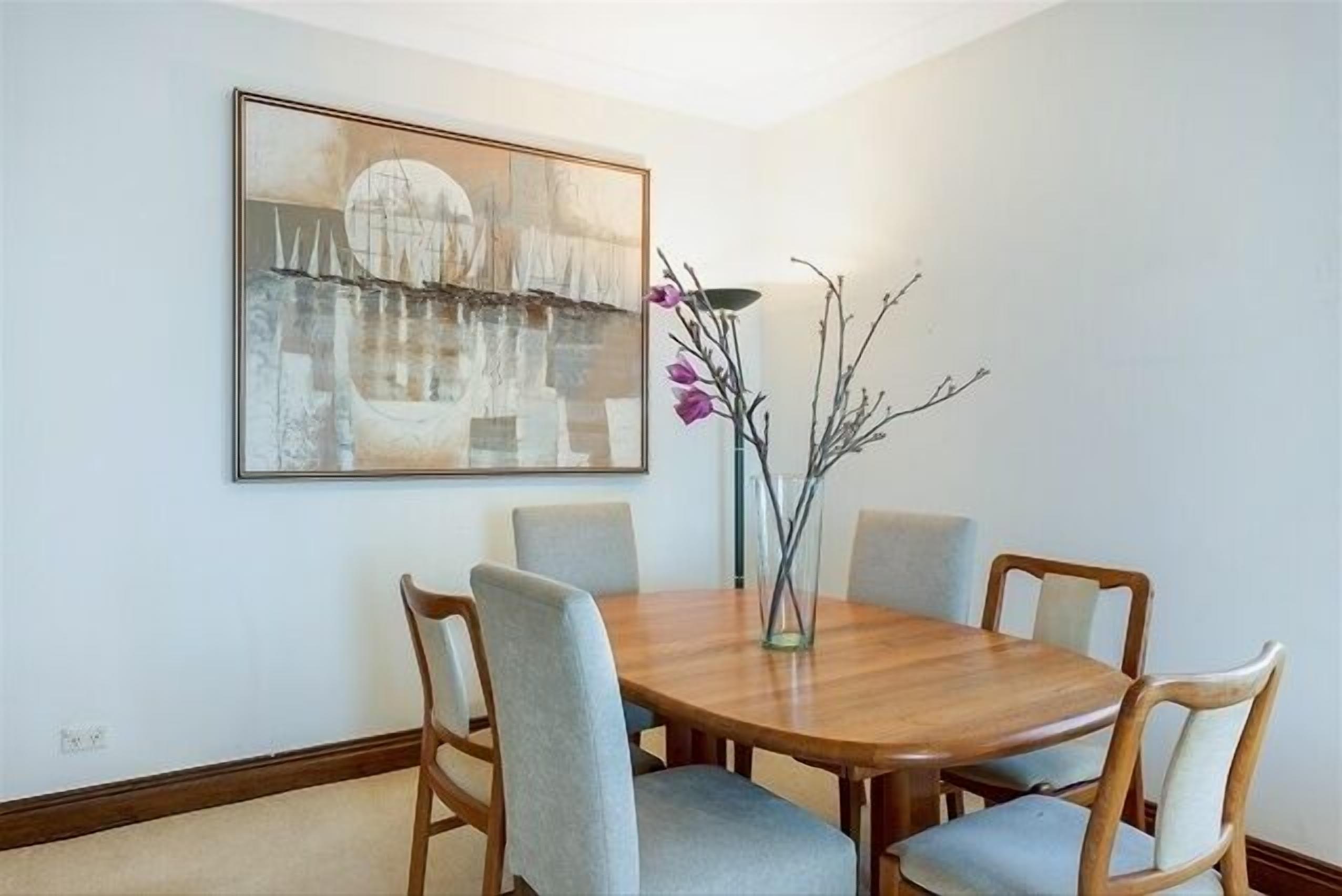 Een eetruimte met een houten tafel en vier stoelen, een vaas met takken, en een groot abstract schilderij aan de muur.
