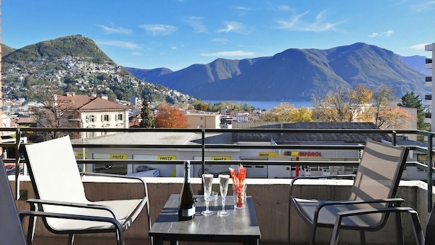 Un balcon avec une table, des chaises, une bouteille et des verres, donnant sur une ville et des montagnes.
