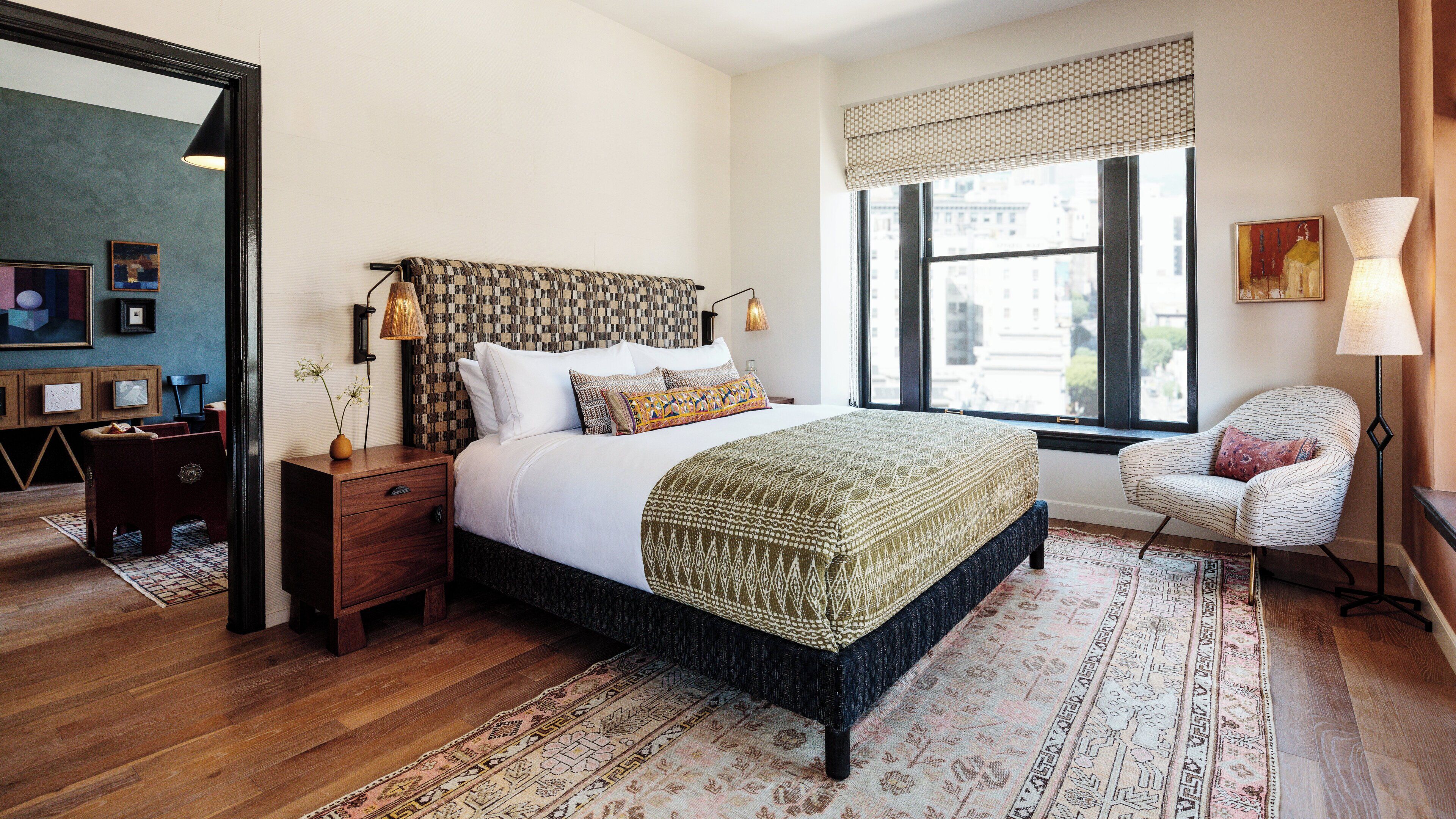 A bedroom with a large bed, a nightstand, a chair, a window with a view of buildings, and a patterned rug.