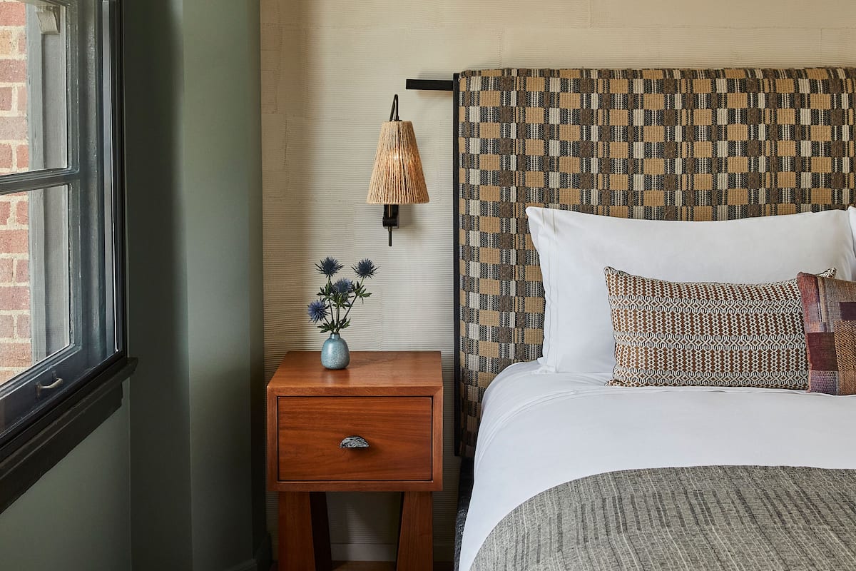 A bedroom with a wooden nightstand, a lamp, a vase with flowers, a patterned headboard, and a bed with white bedding.