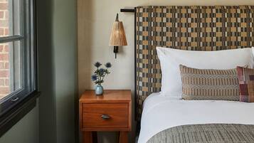 Une chambre à coucher avec une table de chevet en bois, une lampe, un vase de fleurs, une tête de lit à motifs et un lit avec une literie blanche.
