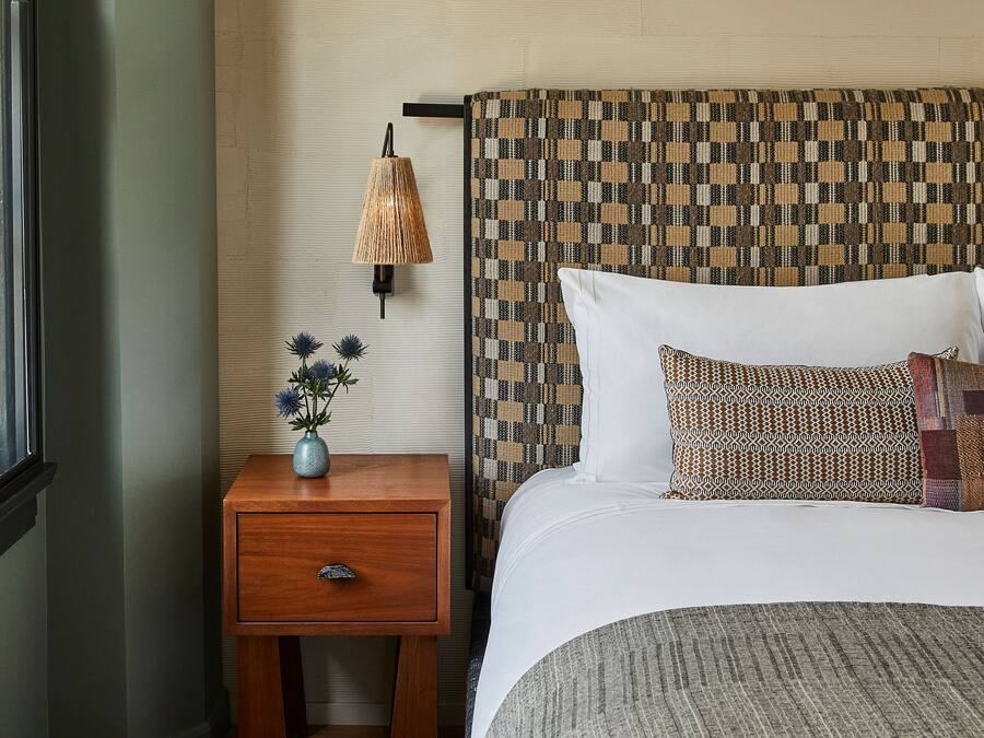 Une chambre à coucher avec une table de chevet en bois, une lampe, un vase de fleurs, une tête de lit à motifs et un lit avec une literie blanche.