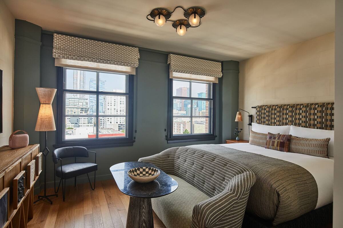 A bedroom with a large bed, two windows with view, a chair, a table with a bowl, and a lamp.