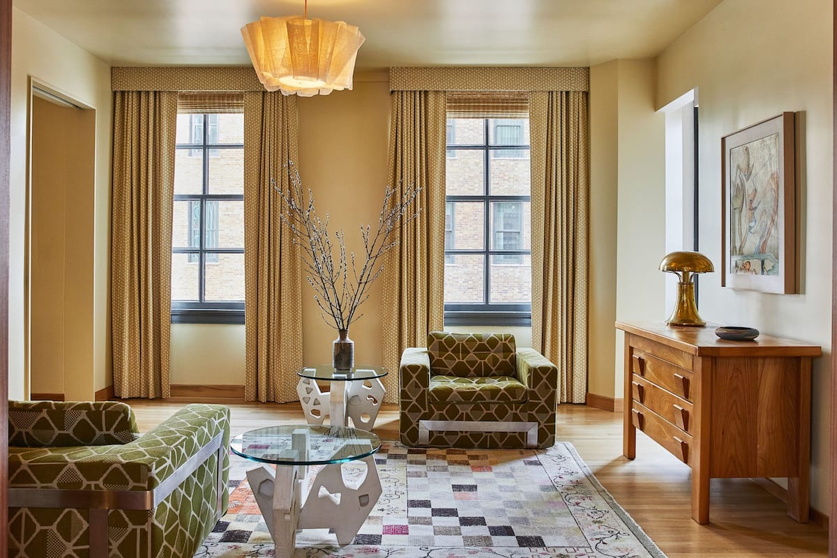 A room with a patterned armchair, a glass coffee table, a vase with branches, a wooden cabinet, and a painting on the wall.