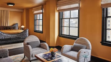 A hotel room with a bed, two armchairs, a coffee table, and large windows with views of buildings.