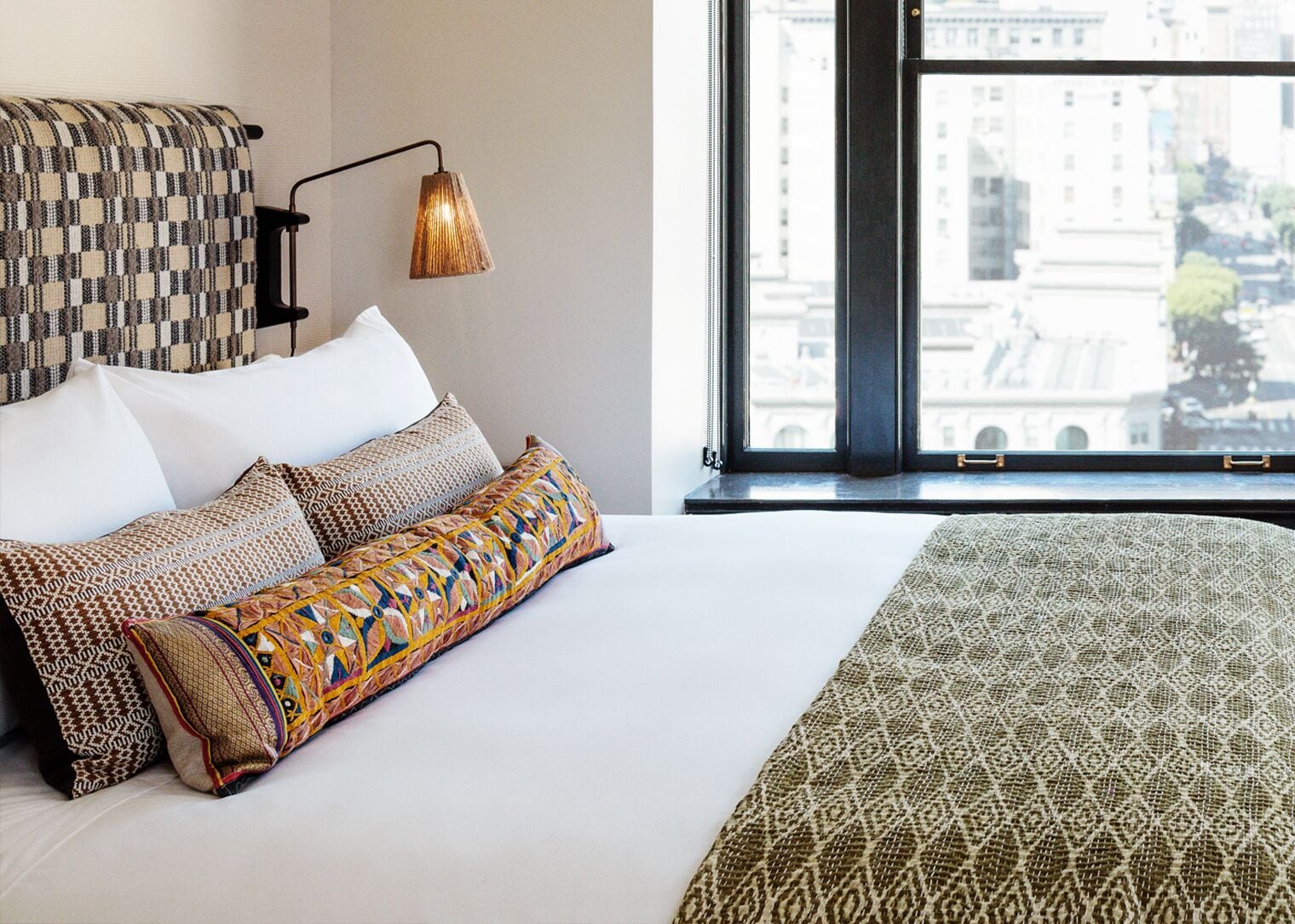 A neatly made bed with patterned pillows and a green blanket, a wall-mounted lamp, and a window with a city view.