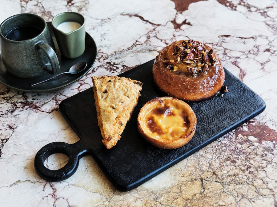 A black serving board with a pastry, a slice of bread, and a cup of coffee on a marble surface.