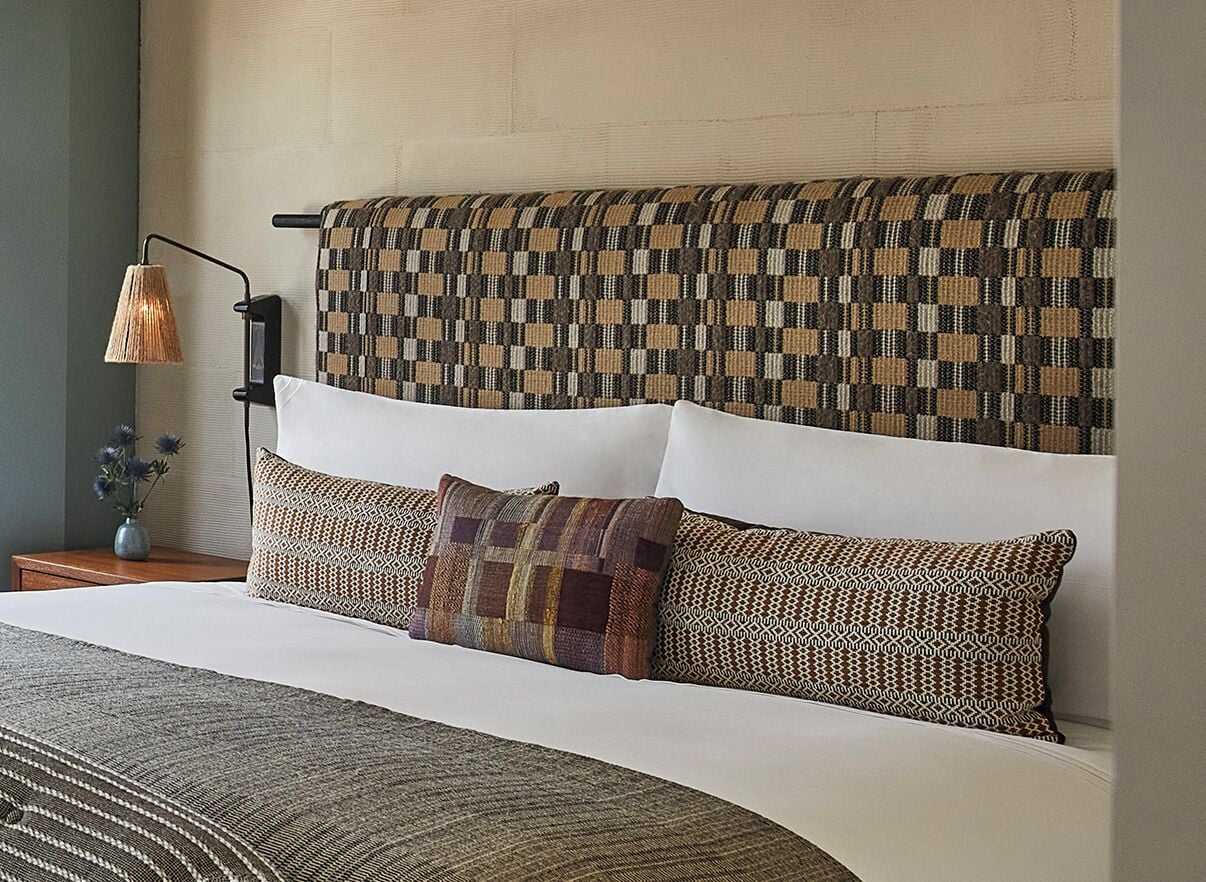 Cama bem arrumada com roupa de cama estampada, abajur e cabeceira fixada na parede.