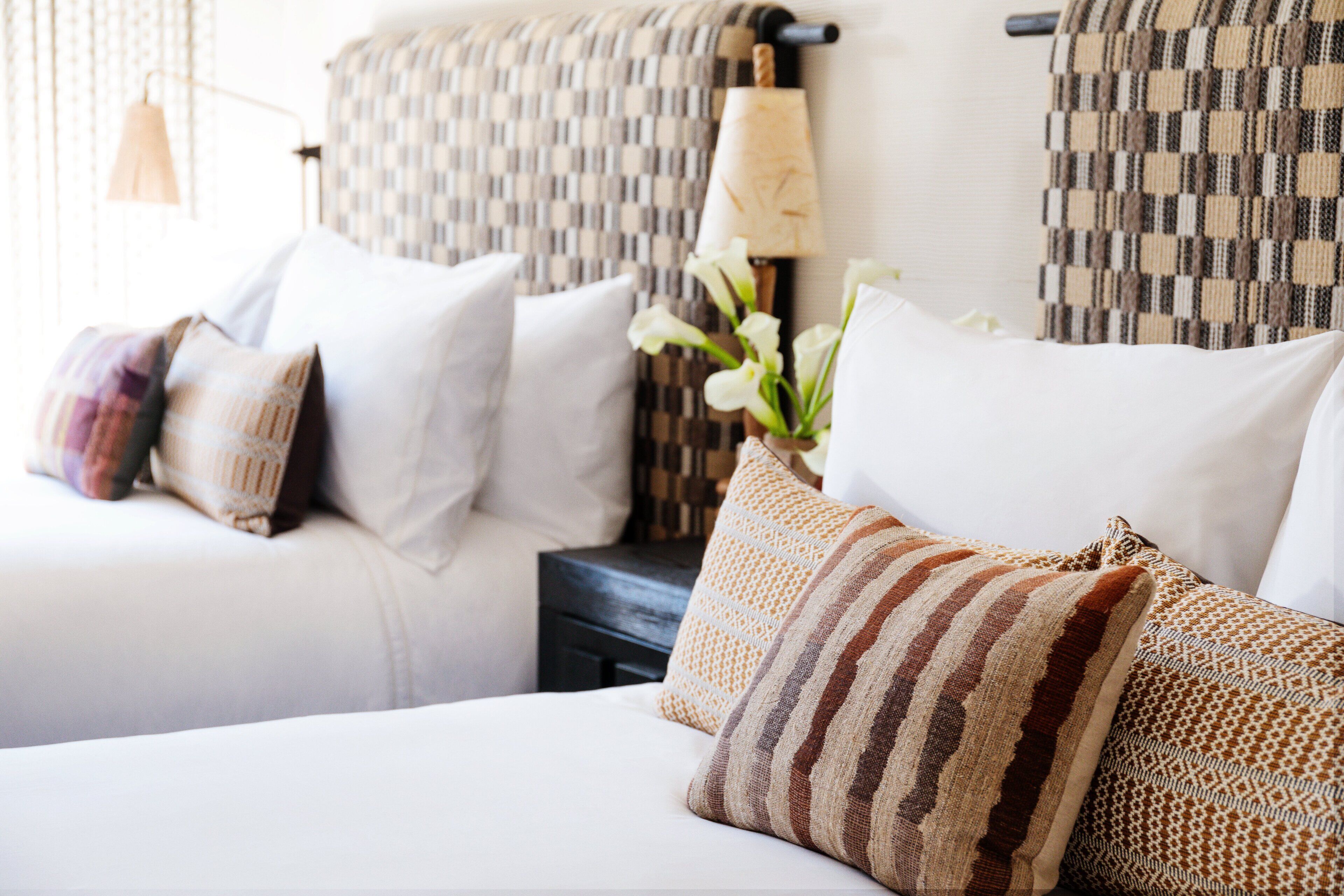 A hotel room with two beds, each with white bedding and multiple pillows. There is a patterned headboard behind the beds, and a small table with a lamp and flowers.