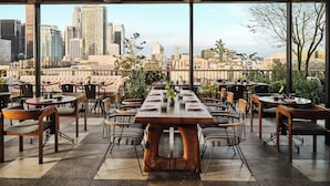 Un espace de restauration en plein air offrant une vue sur les toits de la ville, avec une longue table en bois dressée pour le repas, entourée de chaises et de plantes en pot.