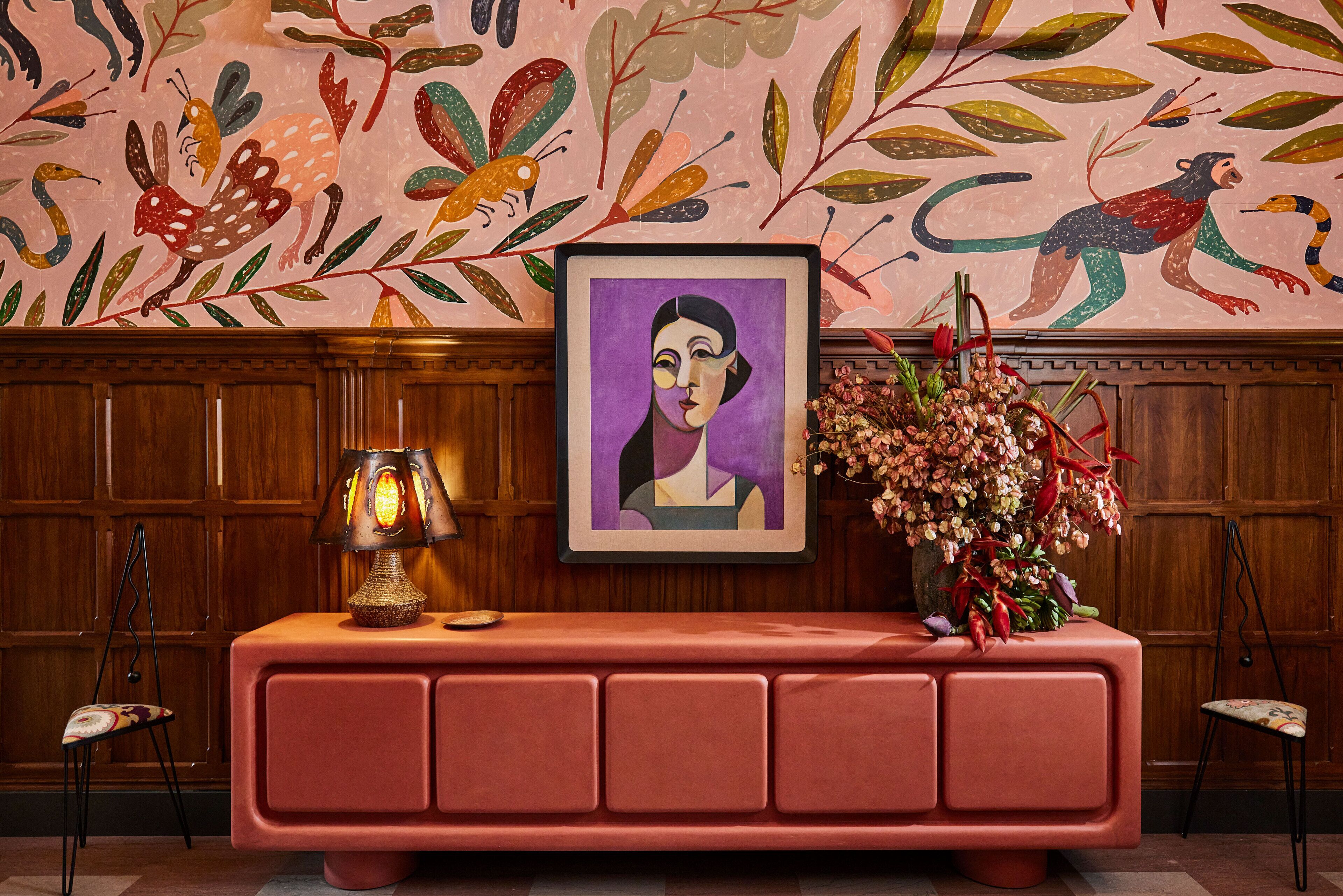 A wooden panel wall with a colorful mural, a framed artwork, a red console table with a lamp, and a vase of flowers.