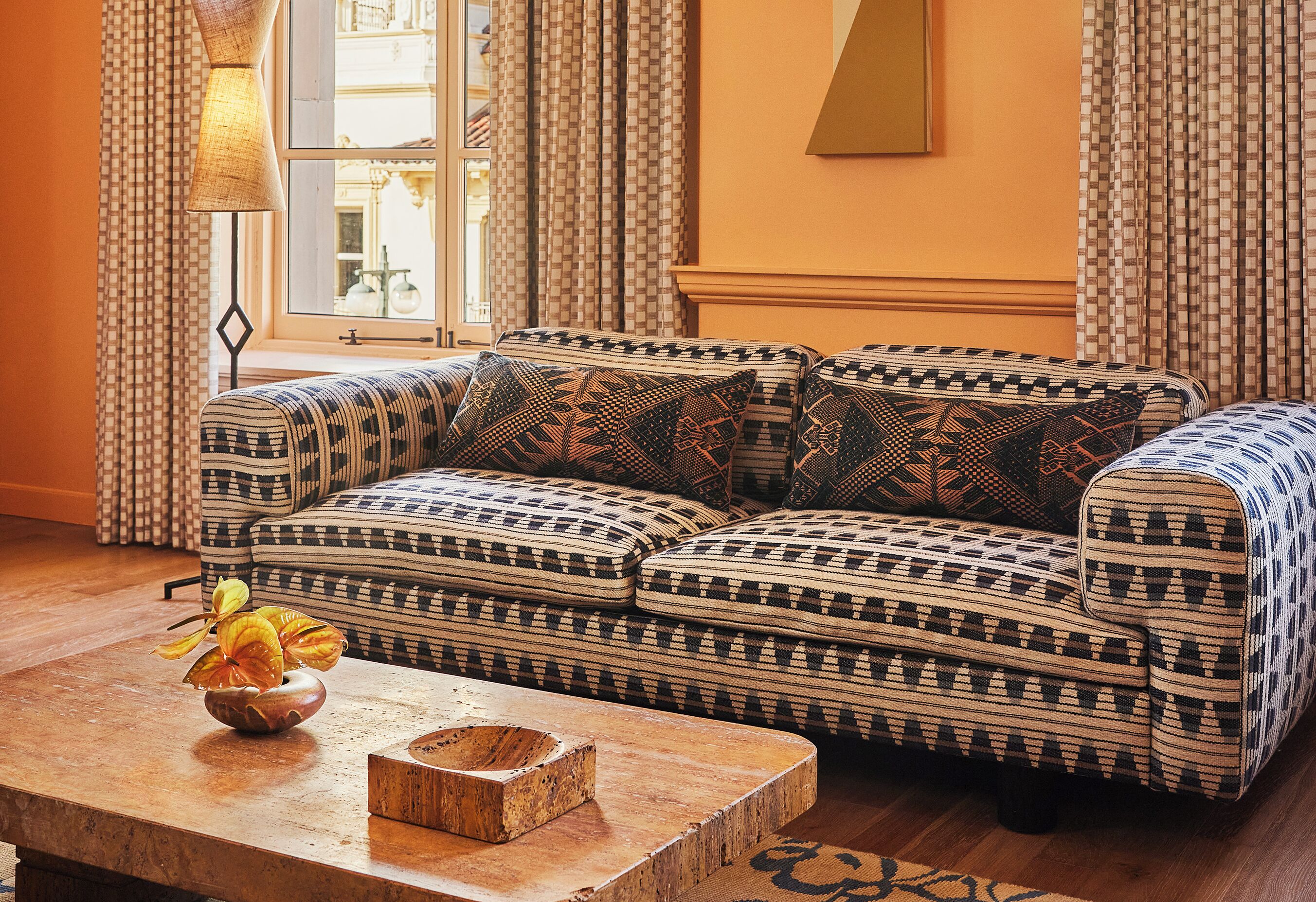 A patterned sofa set in a room with a wooden coffee table, a vase with flowers, and a window with curtains.