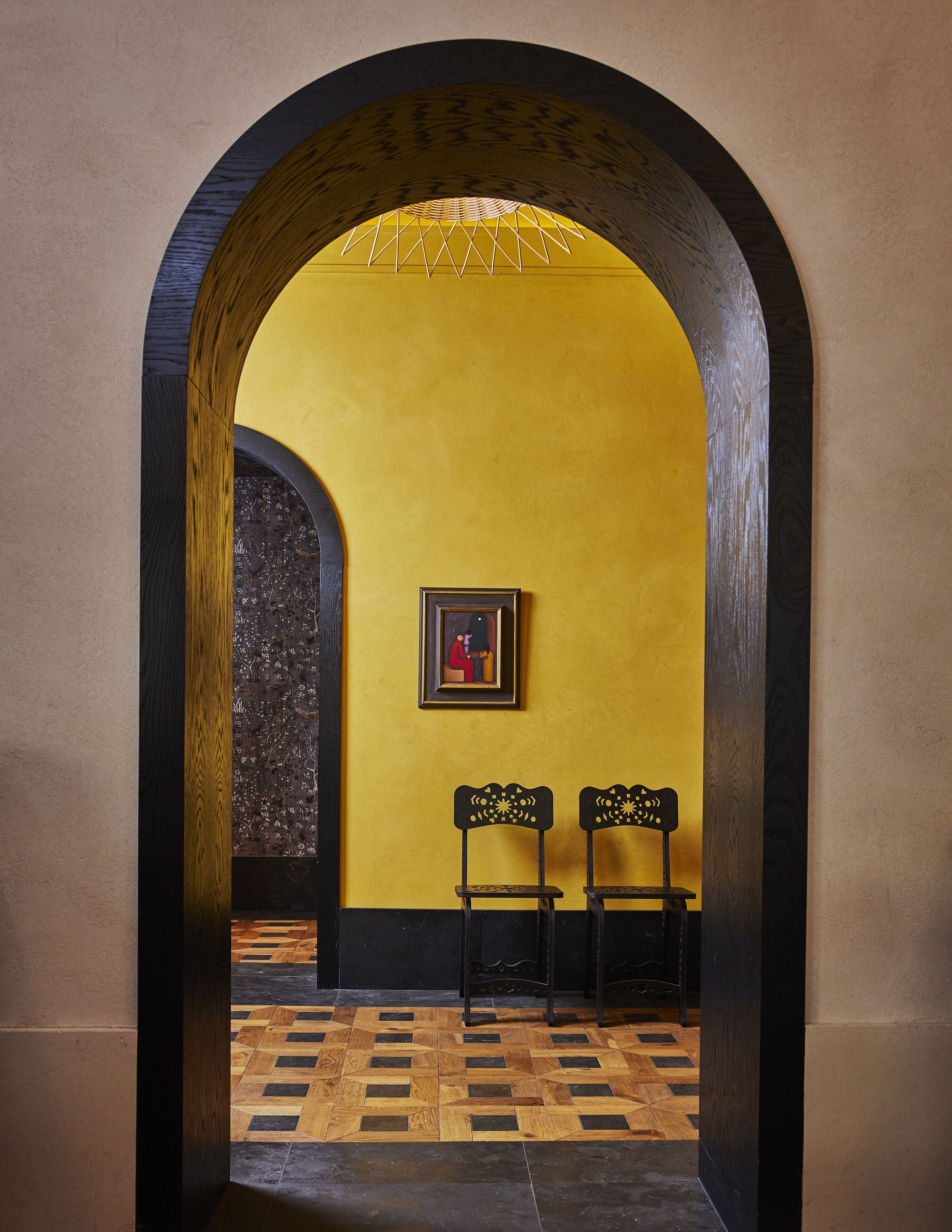 Un arco de entrada conduce a una habitación con una pared amarilla y un cuadro enmarcado. Dos sillas negras están colocadas frente a la pared.