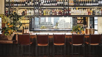A bar with multiple shelves of bottles, a large window with a city view, and several chairs.