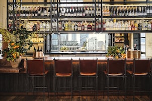 A bar with multiple shelves of bottles, a large window with a city view, and several chairs.