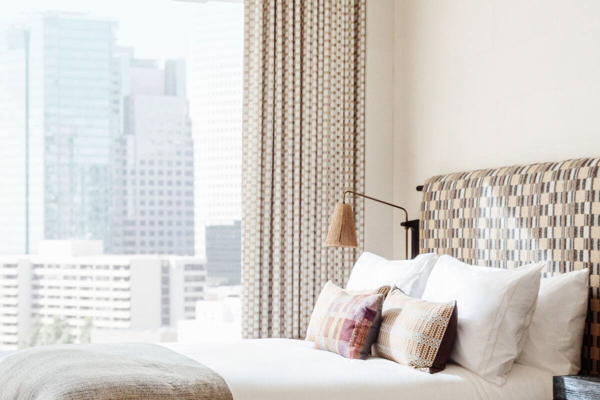 A hotel room with a bed, a window with curtains, and a cityscape view.
