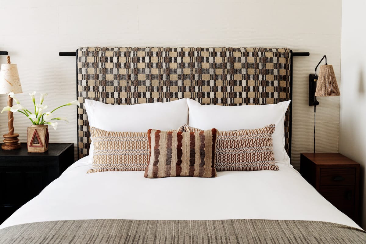 A neatly made bed with a patterned headboard, a bedside lamp, and a small table with a vase and a plant.