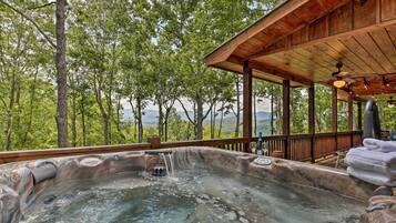 A hot tub with a waterfall feature, surrounded by a wooden deck and overlooking a forested area.